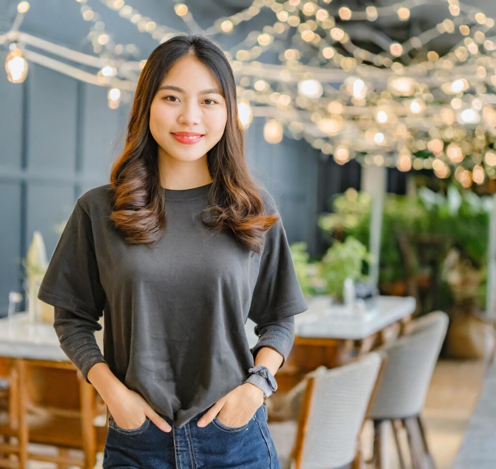 An Asian woman standing in a restaurant adorned with lights. She is wearing the L.L. Bean Women's Carefree Unshrinkable Long Sleeve Tee.