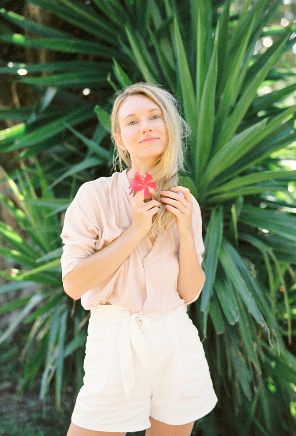 Woman in white linen shorts and pink shirt holding a flower in a sunny garden.
