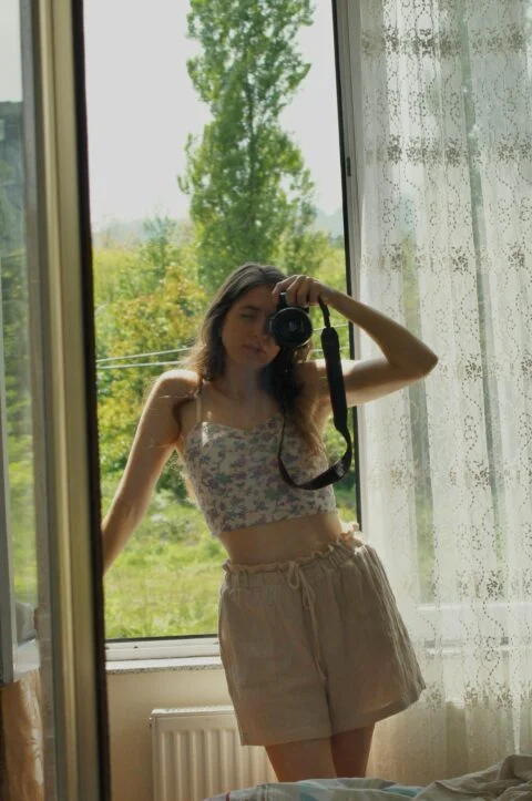 A woman capturing a self-portrait in front of a window, showcasing her style while wearing linen shorts