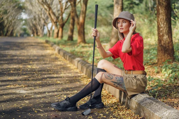 A person wearing a red shirt, linen shorts, and a hat sits on the curb of a tree-lined path, holding a golf club. They have a thoughtful expression and visible tattoos on their leg. Dry leaves scatter the ground, hinting at autumn.