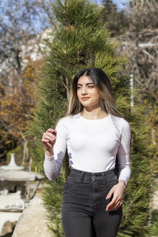 A stylish woman wearing a fitted white Henley shirt and jeans poses in front of a vibrant tree, showcasing her casual look.