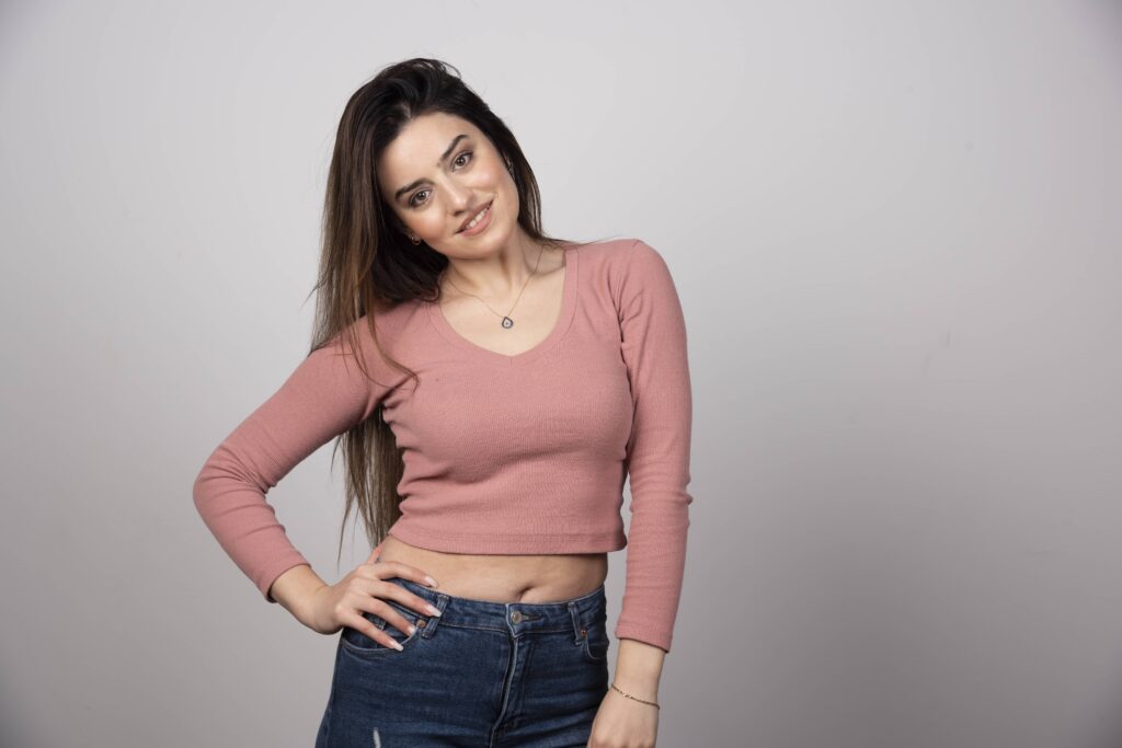 A beautiful young woman wearing a pink cropped long sleeve t-shirt and jeans with long hair standing with hand on her waist.