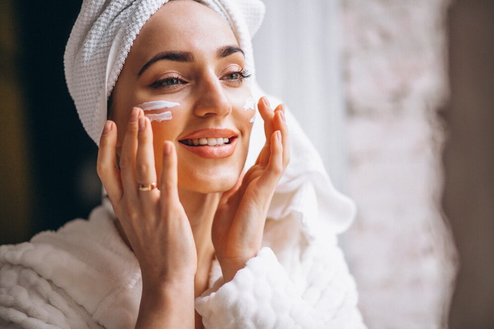 A young woman with a towel wrapped around her hair, smiling as she applies acne cream to her face