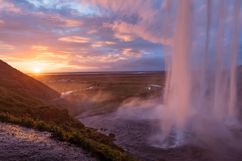 Seljalandsfoss waterfall captured during sunset, with the sun setting behind distant mountains and warm hues painting the sky.