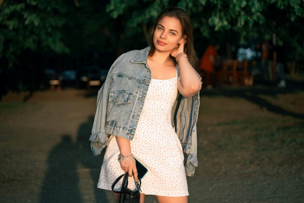A young woman in a white slip dress with small prints, layered with a denim jacket, enjoying a casual day out in a park.