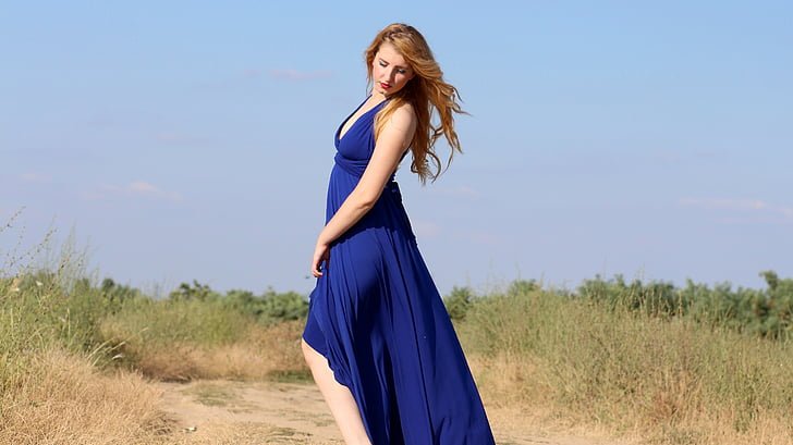 A woman in a flowing blue cotton slip dress stands on a dirt path in a rural area