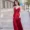 A woman in a vibrant red slip dress is walking down a city street. She has shoulder-length brown hair and is smiling gently while looking slightly to her left.