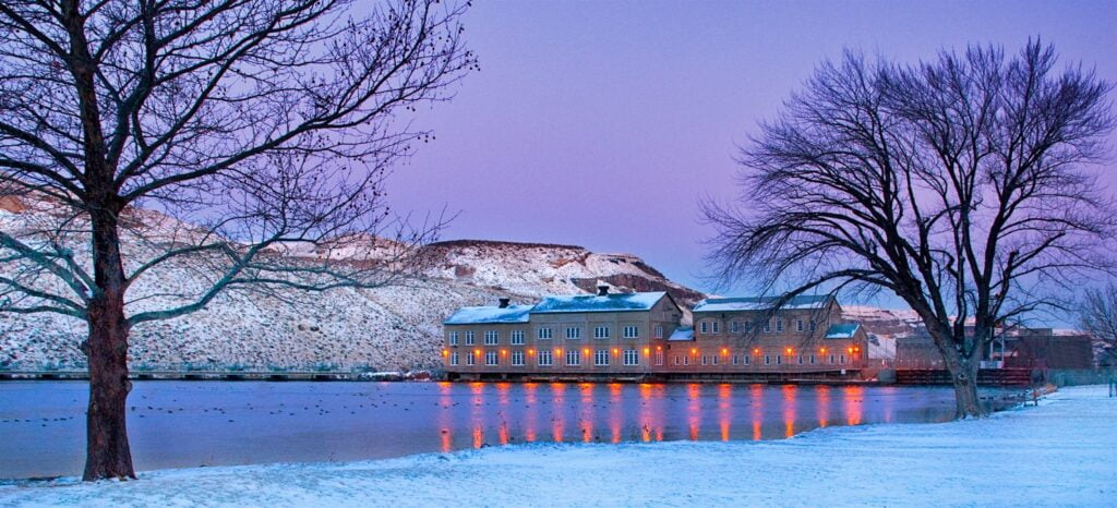 The Swan Falls Dam in Idaho, surrounded by snow-covered ground and trees, is illuminated by warm lights reflecting off the calm water at dusk.