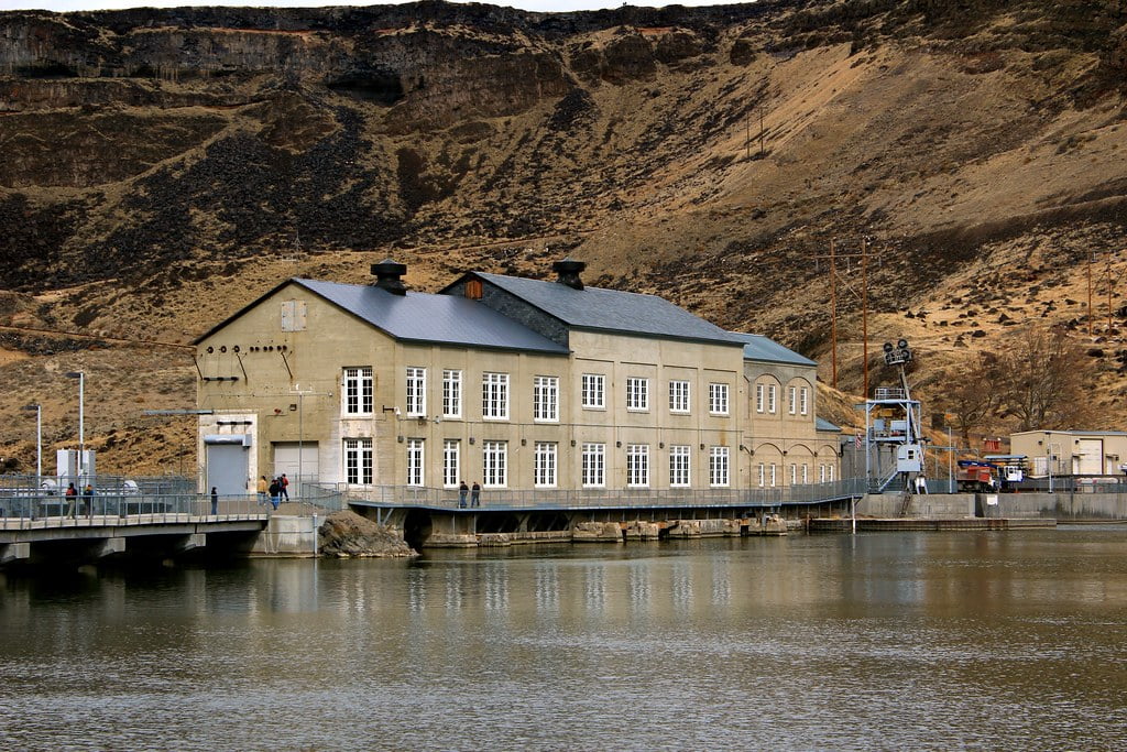 A historic building of the Idaho Power Company, located by the water with rugged hills in the background, features large windows and a bridge with people walking.