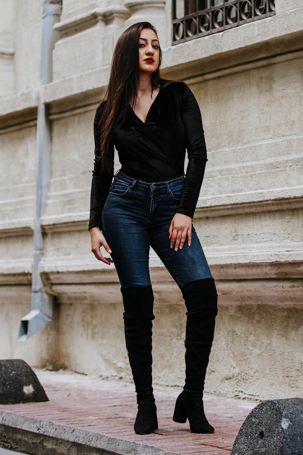 A woman with long dark hair stands against a beige wall. She is wearing a black long-sleeved top, wide-leg jeans, and black ankle boots. She looks confidently at the camera, with one hand resting on her thigh and the other hanging by her side.
