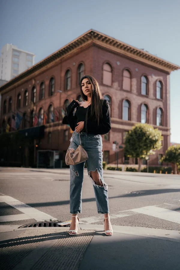 A woman stands confidently at a street corner in front of a large brick building. She wears a black top, wide-leg jeans, and white sandals, carrying a beige handbag. The sky is clear, and trees and flags are visible around the building.