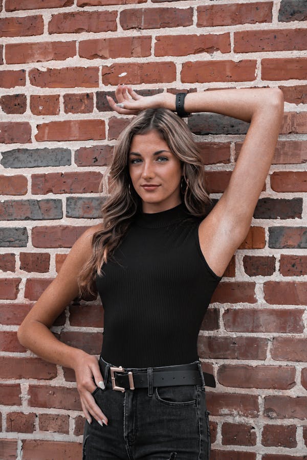 A woman with long, wavy brown hair poses confidently against a brick wall. She is wearing a black sleeveless top and wide-leg jeans with belts, with one arm resting on her head and the other hand placed on her hip, showcasing a modern and stylish look.