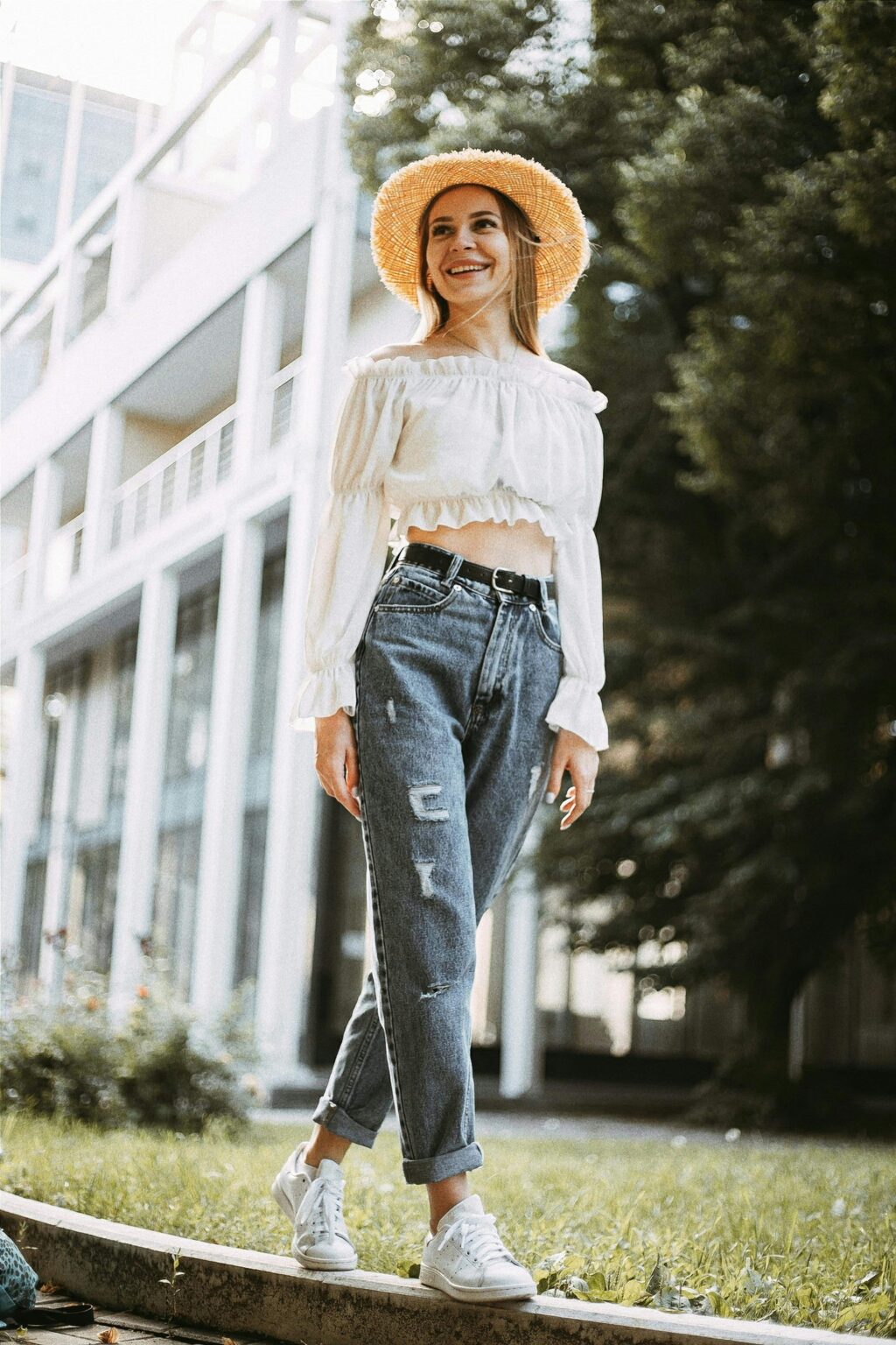 A woman wearing a straw hat, white blouse, and high-waisted wide-leg jeans stands outdoors. She smiles and looks into the distance, with trees and a modern building in the background.