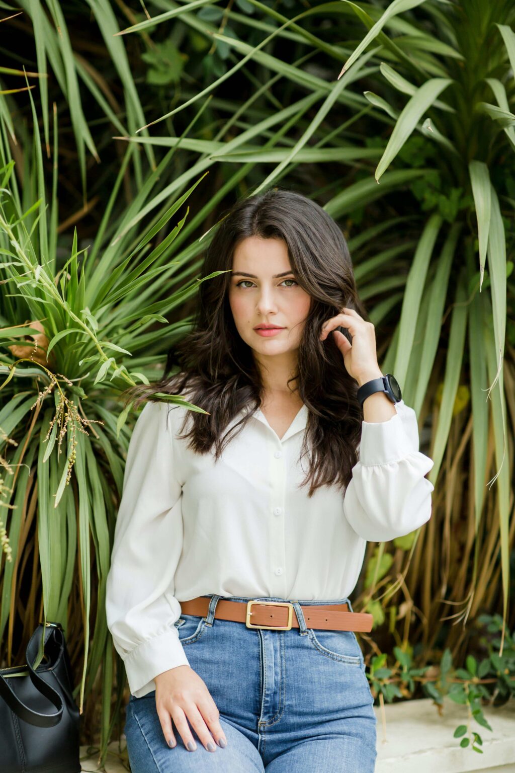 A woman with long dark hair, wearing a white Button-Down Shirt that's tucked into wide-leg jeans with a brown belt, stands in front of lush green foliage. She holds one hand near her face while the other rests on her thigh.