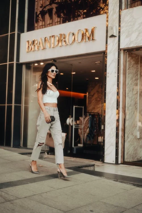 A woman wearing a white top, wide-leg jeans with heels, and sunglasses is walking in front of a store named "BRANDROOM." She is carrying a handbag and the store displays clothes inside. The background features glass windows and a marble storefront.