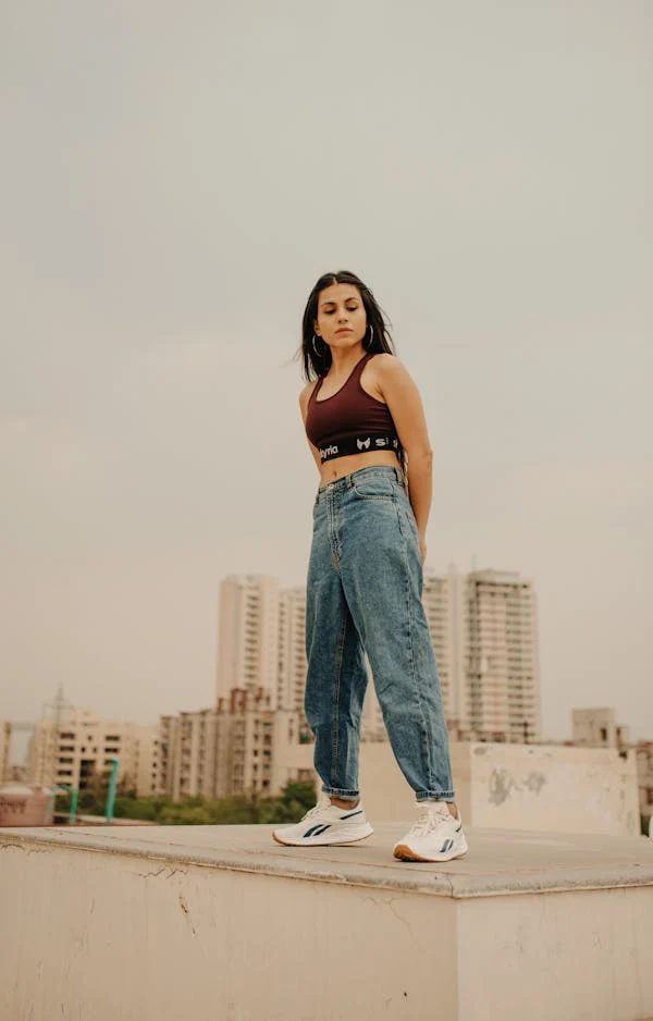 A woman stands confidently on a rooftop, wearing a maroon sports bra, wide-leg jeans with sneakers. She gazes downward with her hands behind her back, with a cityscape of tall buildings in the background under a cloudy sky.