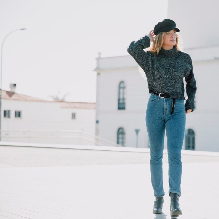 A woman is standing outdoors in a bright, spacious area with a white building in the background. She is wearing a black hat, a dark high-neck sweatshirt, wide-leg jeans, and black ankle boots, with one hand on her hat and the other resting by her side.