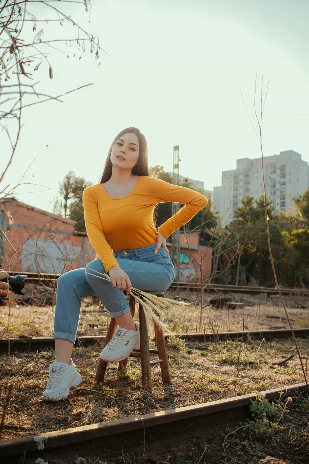 A person with long hair wearing a yellow T-shirt, wide-leg jeans, and white sneakers sits on a wooden stool outdoors. They are posed with one hand on their hip and the other resting on the stool, with a cityscape and trees in the background.