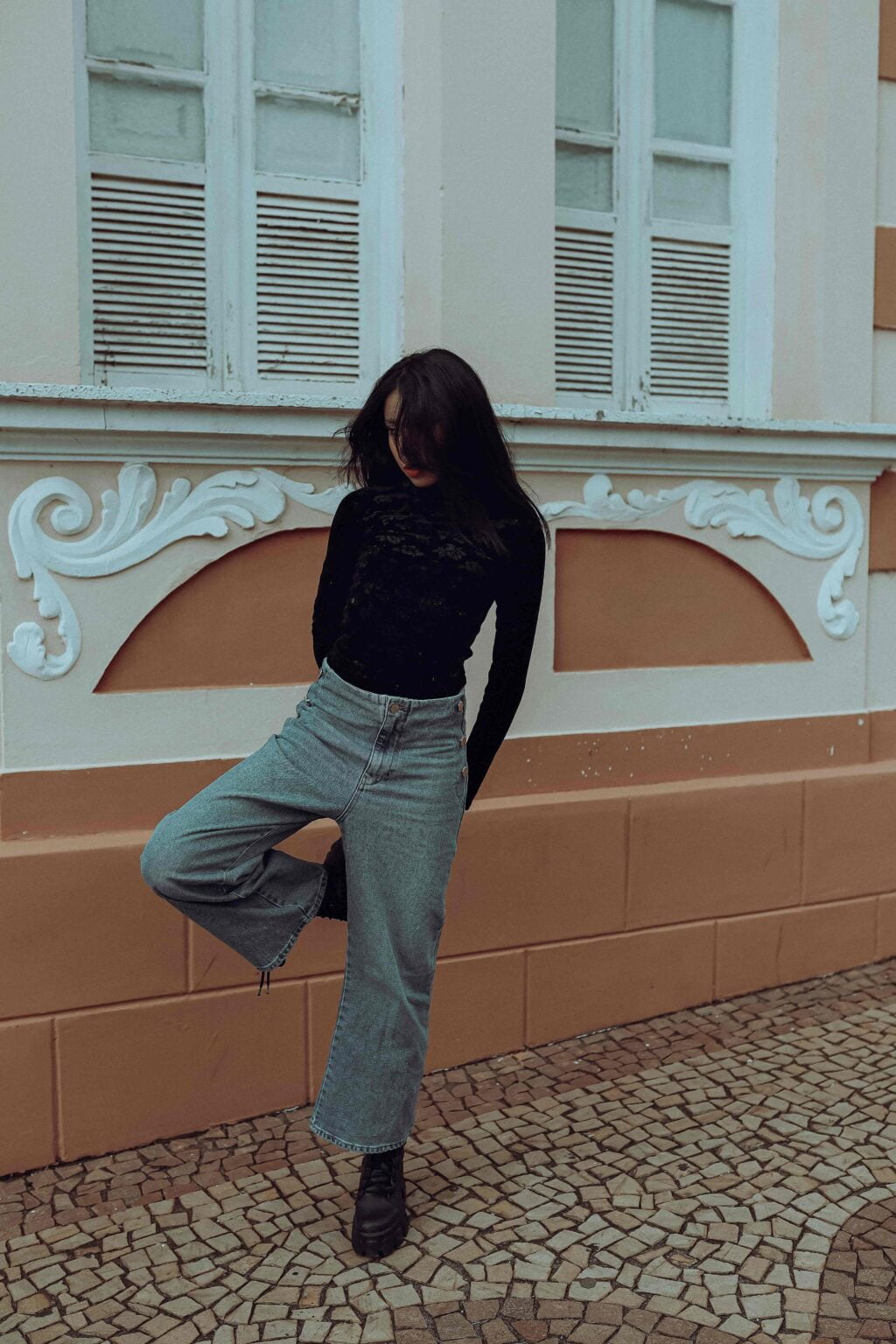 A Women with long dark hair stands in front of an ornate building with shuttered windows. They are wearing a black fitted turtleneck and high-waisted wide-leg blue jeans, with one leg slightly raised and hands behind their back. The ground is covered in patterned tiles.