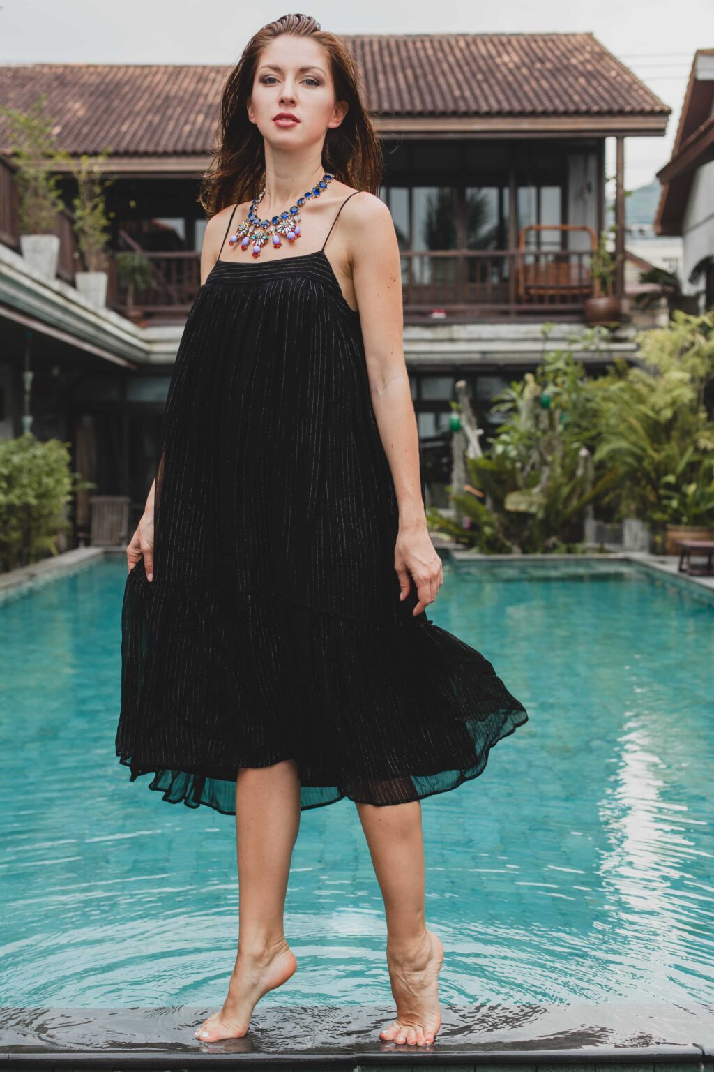 A woman in a loose fit slip dress stands barefoot by a pool, with a confident expression and a statement necklace, in front of a villa.
