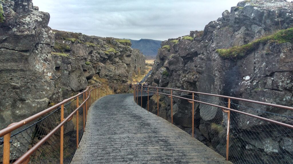A wooden walkway with handrails on either side winds through a dramatic landscape of rugged, rocky cliffs, tracing the tectonic plate boundaries at Thingvellir National Park in Iceland. The path leads toward distant mountains beneath a cloudy sky, surrounded by patches of moss and sparse vegetation.