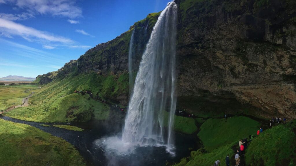 People are walking behind the Seljalandsfoss in Iceland, where the waterfall cascades down a mossy cliff into a pool below, with a vast green landscape and a blue sky in the background