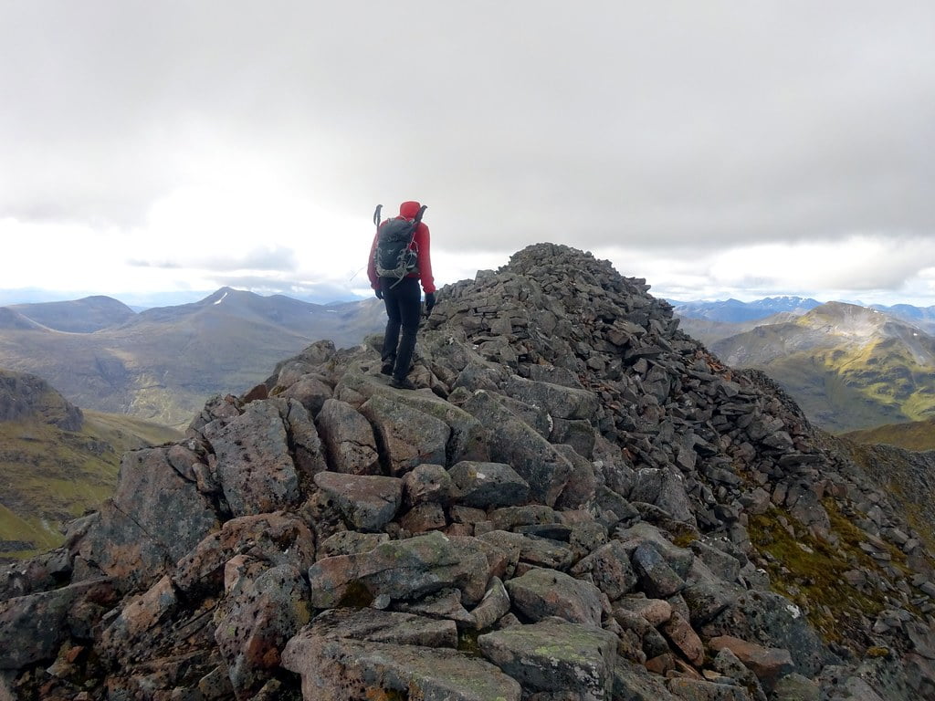 A person with a backpack stands at the peak of Ben Nevis, enjoying the stunning panoramic views from the mountain summit.
