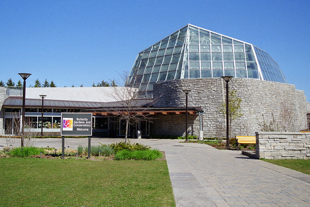 A modern, glass-domed Butterfly Conservatory Niagara Falls with a stone facade, surrounded by a neatly landscaped garden and a sign that reads 'Butterfly Gardens and Conservatory Welcome.' The conservatory is bathed in sunlight under a clear blue sky."