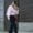 "A woman walks confidently on a paved sidewalk, dressed in a light pink blouse with puffed sleeves, dark velvet pants, and colorful platform sandals. She accessorizes with a patterned scarf around her neck and carries a small red purse."