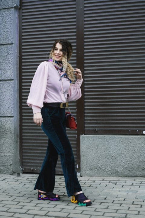 "A woman walks confidently on a paved sidewalk, dressed in a light pink blouse with puffed sleeves, dark velvet pants, and colorful platform sandals. She accessorizes with a patterned scarf around her neck and carries a small red purse."