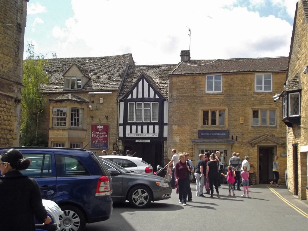 People stroll along the street by Cotswolds Perfumery, with parked cars lining the picturesque road in Bourton-on-the-Water.