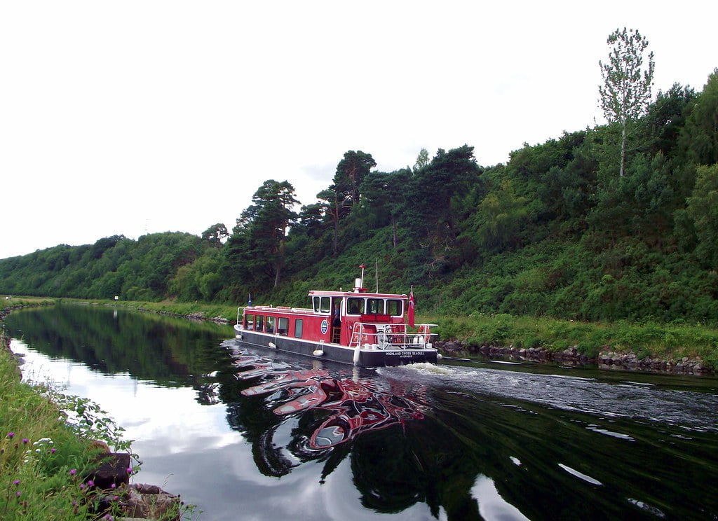 A red and white boat is cruising along a calm canal surrounded by lush green trees and foliage. The boat's reflection is clearly visible in the still water. The sky is overcast, adding a serene ambiance to the picturesque scene, reminiscent of a Cruise Loch Ness through Loch Linnhe.