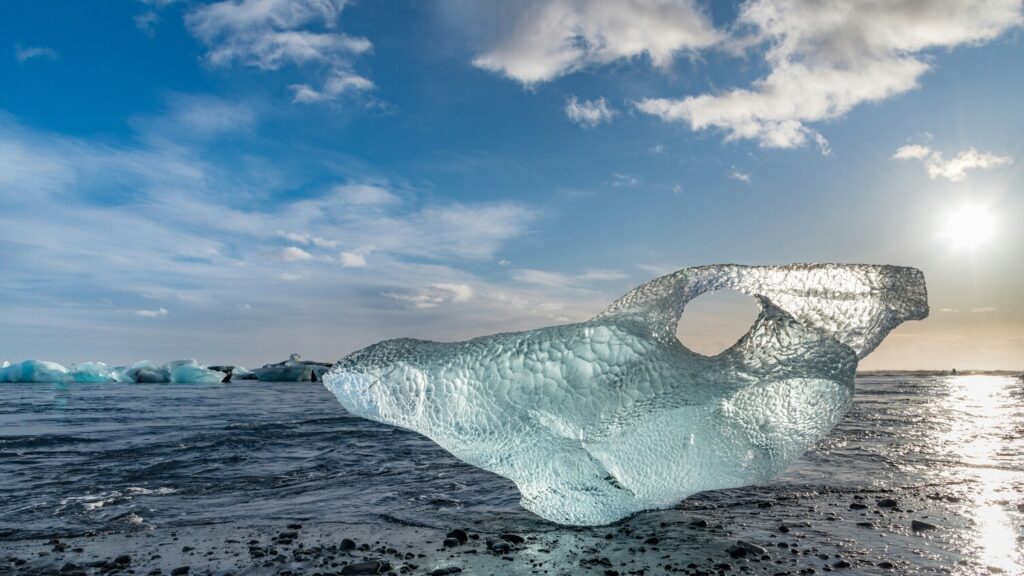 A large, sculpted iceberg with a hole in it rests on Diamond Beach in Iceland. The sun is shining brightly in the clear blue sky, reflecting off the calm sea and the icebergs scattered in the background.