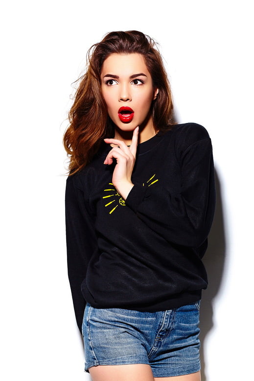 A woman poses against a white background, wearing a black long sleeve round neck casual T-shirt and denim shorts. Her hair is styled in loose waves, and she sports a bright red lipstick, with a surprised expression on her face.