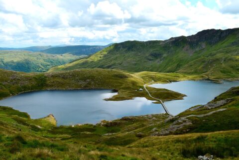 Snowdonia National Park landscape with two tranquil lakes linked by a slender pathway. The scene is enveloped by lush, green hills, with mountains visible in the background beneath a sky dotted with clouds.
