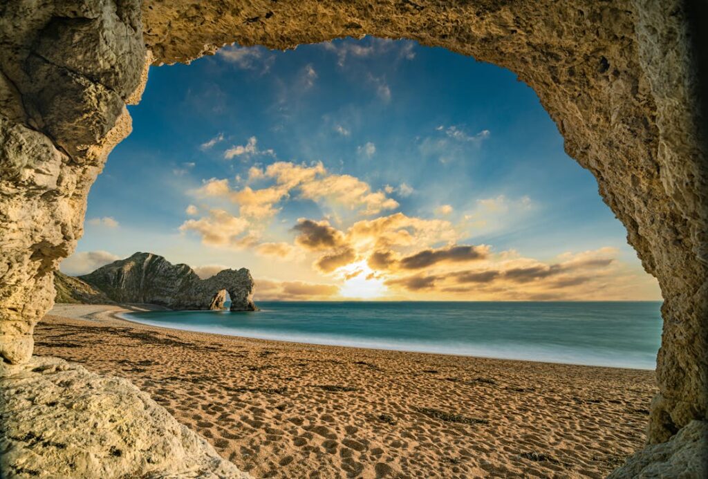 A breathtaking view of the Jurassic Coast at sunset, seen through a rocky archway framing the sandy beach, calm sea, and dramatic cliffs under a vibrant sky with golden clouds.