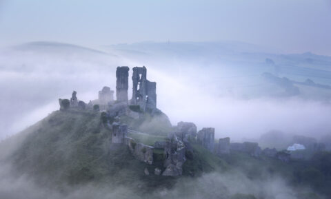 A foggy scene featuring Corfe Castle, with its ancient stone walls partially obscured, evoking a sense of mystery and history.