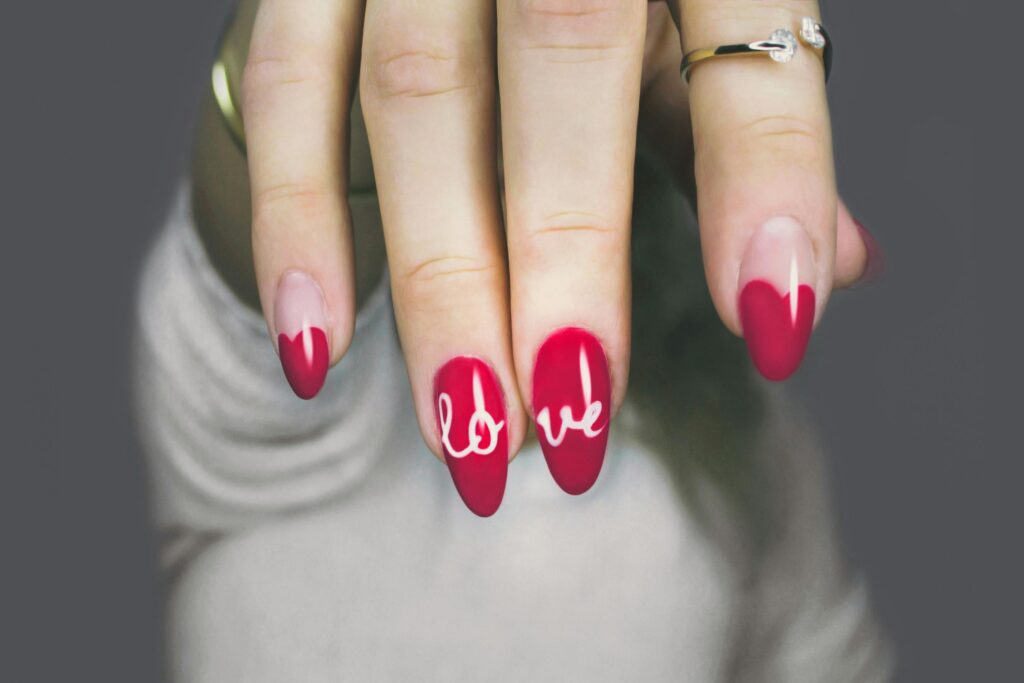 A close-up of a hand with red almond-shaped nails. The word "LOVE" is written in white cursive lettering across the nails.
