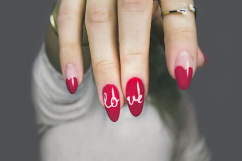 A close-up of a hand with red almond-shaped nails. The word "LOVE" is written in white cursive lettering across the nails.