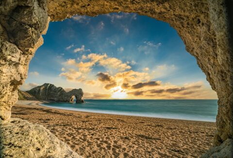 A breathtaking view of the Jurassic Coast at sunset, seen through a rocky archway framing the sandy beach, calm sea, and dramatic cliffs under a vibrant sky with golden clouds.
