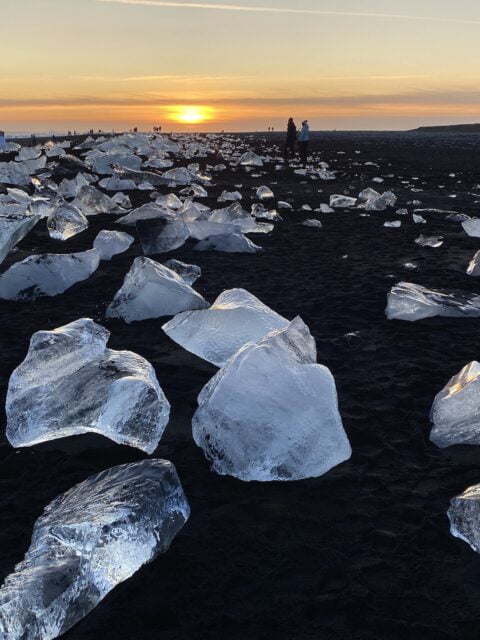 Large chunks of ice scattered across the black sand of Diamond Beach in Iceland, with a sunset in the background. Several people are walking and admiring the scene.