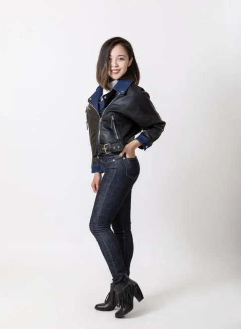 A woman wearing a black leather jacket, dark skinny jeans, and black ankle boots with fringe, standing and smiling against a plain white background.