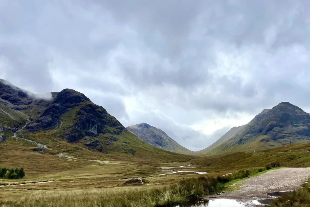 A scenic landscape of Glen Nevis, a lush green valley surrounded by towering mountains. The sky is overcast with dense clouds, casting a serene, moody atmosphere on the scene.