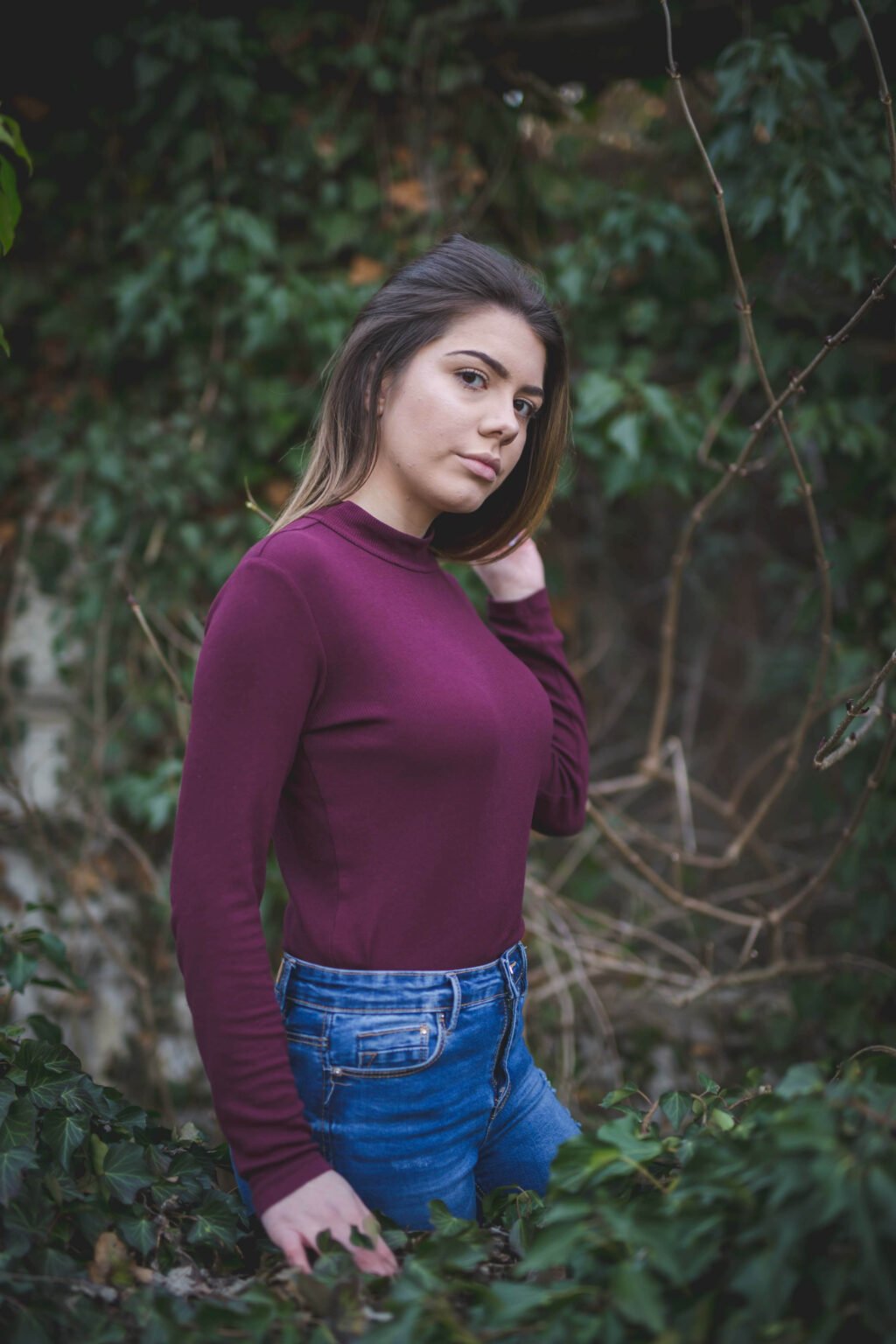 In a tranquil forest, a woman wearing jeans and a maroon high neck long sleeve shirt enjoys the beauty of nature around her.