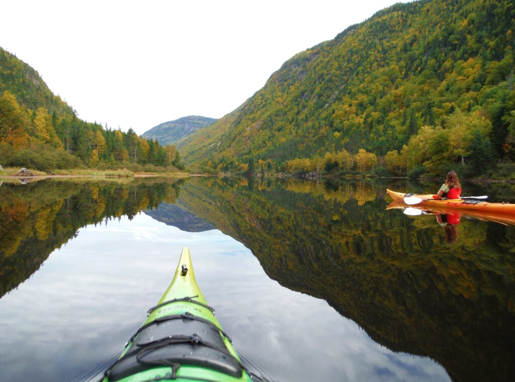 A view from a green kayak on a calm lake surrounded by forested mountains. Another kayaker in an orange kayak is paddling on the right. The trees have autumn colors, and the water reflects the surrounding landscape and the clear sky, reminiscent of kayaking through Loch Linnhe's serene waters.