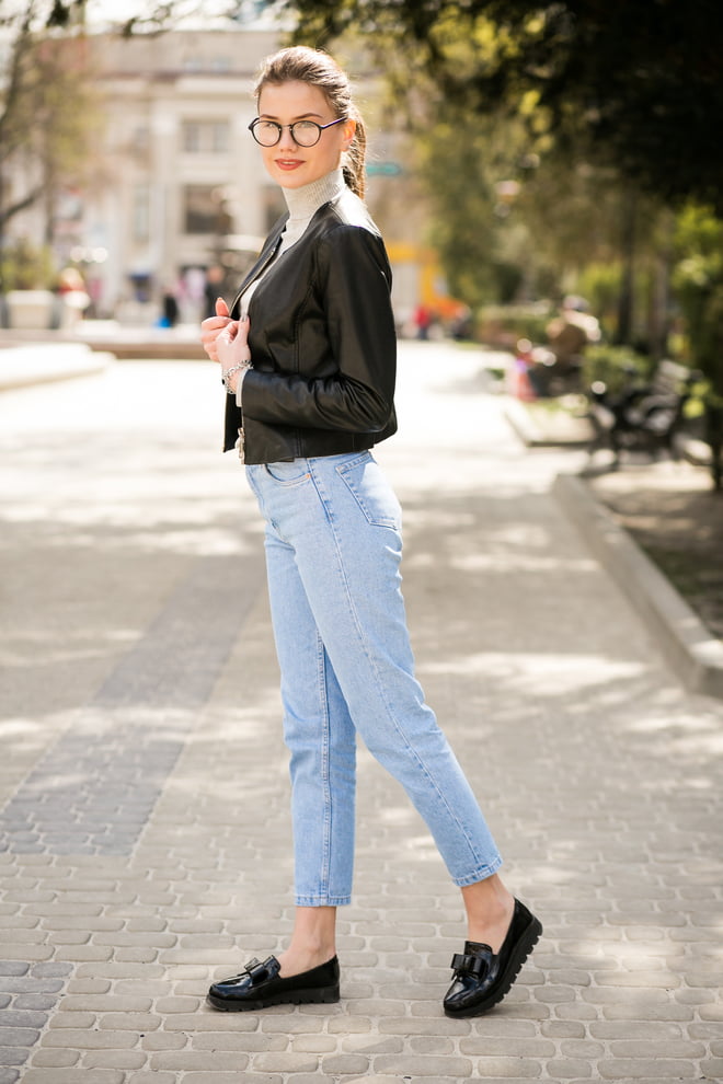 A woman wearing glasses and a black leather jacket, paired with skinny jeans and loafers, exuding a stylish vibe.