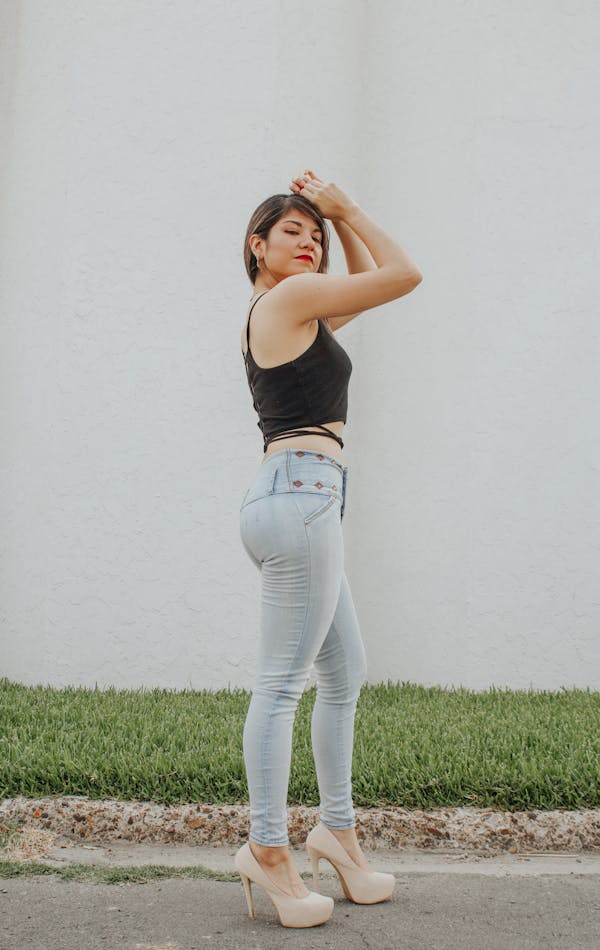 A woman in light blue skinny jeans, a black sleeveless crop top, and beige high heels, posing outdoors against a plain white wall with her arms raised above her head.