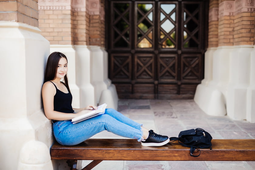A young woman in skinny jeans and sneakers sits on a bench, resting her backpack beside her, enjoying a moment of relaxation.