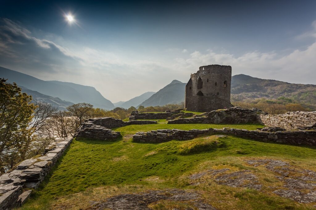 A sunlit scene in Snowdonia National Park featuring the ruins of an ancient stone structure on a grassy hill. The ruins overlook a valley with misty mountains in the background under a bright, hazy sky.