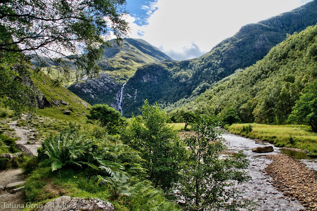 A serene river flows through a lush valley, framed by majestic mountains near Steall Falls, creating a picturesque landscape.
